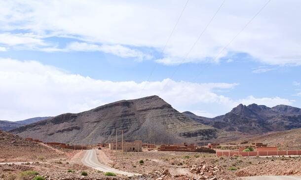 A dirt road in a desert passes a series of flat buildings, with mountains in the background.