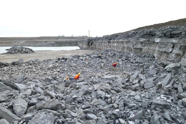 Wide shot of two people in hard hats and bright orange vests crouched separately among the broken grey stones of a wide flat quarry pit and dwarfed by their surroundings.