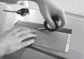 Two hands covering a small rectangular piece of cardboard with black electrical tape, using a tabletop for support. A pair of scissors are also on the table.