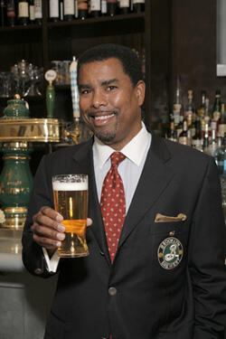 A smiling man, Garrett Oliver, Brooklyn Brewery Brewmaster, posing for the camera while holding a glass of beer. A bar with bottles of spirits is in the background.