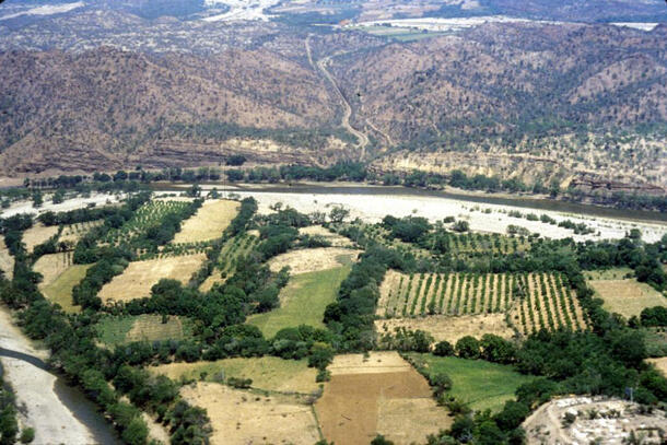 A wide aerial view of an arid environment with some areas with rows of cultivated vegetation. In the foreground is an excavation site. In the distance is a narrow pass through arid scrubby hills.