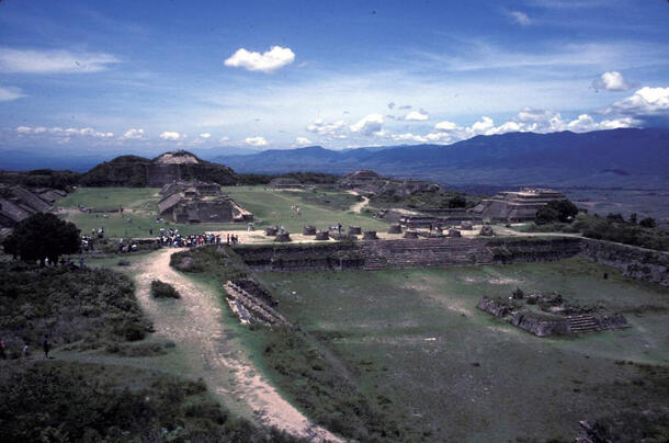 The main plaza at Monte Alban, showing excavated building structures on a wide flat grass-covered land area beneath a clear blue sky, ringed with hills in the distance.