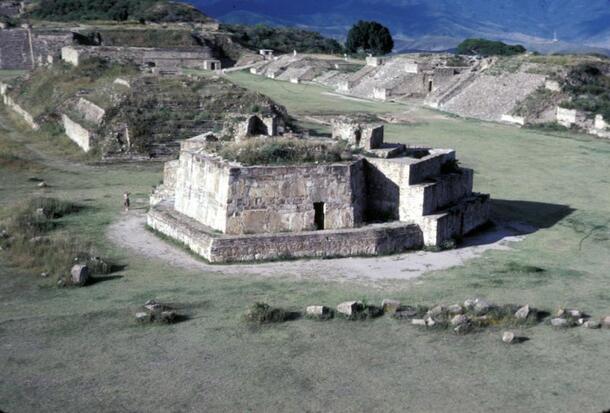 A stone building among other stone structures on a flat plaza with sparse dry grass.