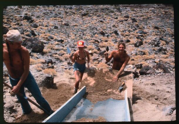 Young people with trowels sifting dirt that is coming down a slide in an arid outdoor location.