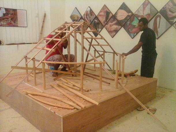 Two people working on a model of the Museum of Samoa. Currently it appears as a frame of wood poles on a wooden platform.