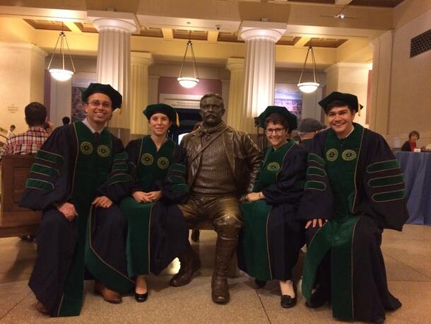 Four PhD graduates wearing their caps and gowns sit beside the Theodore Roosevelt statue in the Theodore Roosevelt Memorial Hall.