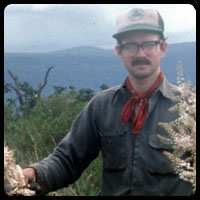 Man posing for photo outdoors in a hilly area, holding plants.