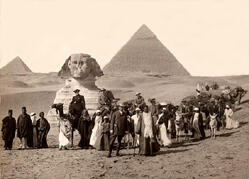 A large group of expedition members dressed in light and dark robes stand before a sphinx and pyramids in the Fayum Basin of Egypt, in 1902.