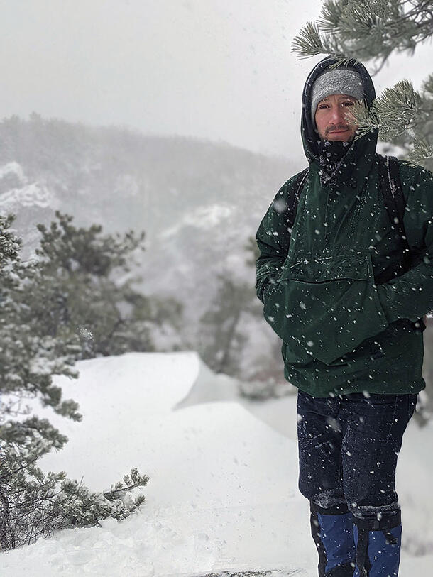 Jesse Delia wears a winter hat and coat and stands with his hands in his pockets in front of snow-covered trees as snow falls around him.