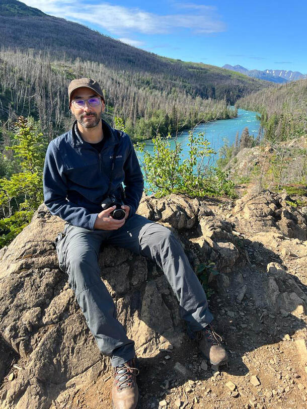 Marcus Brandao is seated on a large rock and holds a camera, with a river, trees, and a distant mountain in view in the background.