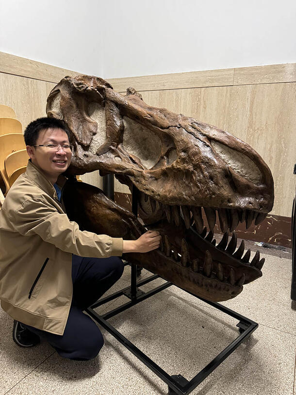 Yulin Yu kneels next to a huge dinosaur skull that is mounted on a metal bracket.