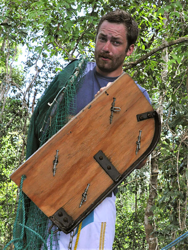 Maxwell Bernt stands amongst trees and holds a wooden board with his left hand and a net with his right hand.