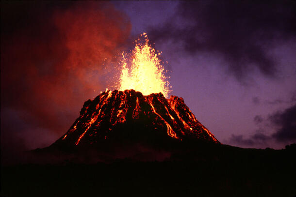 Photo of an erupting volcano shows fire spewing up into a dark sky and lava rolling in streams down the sides of the cone.