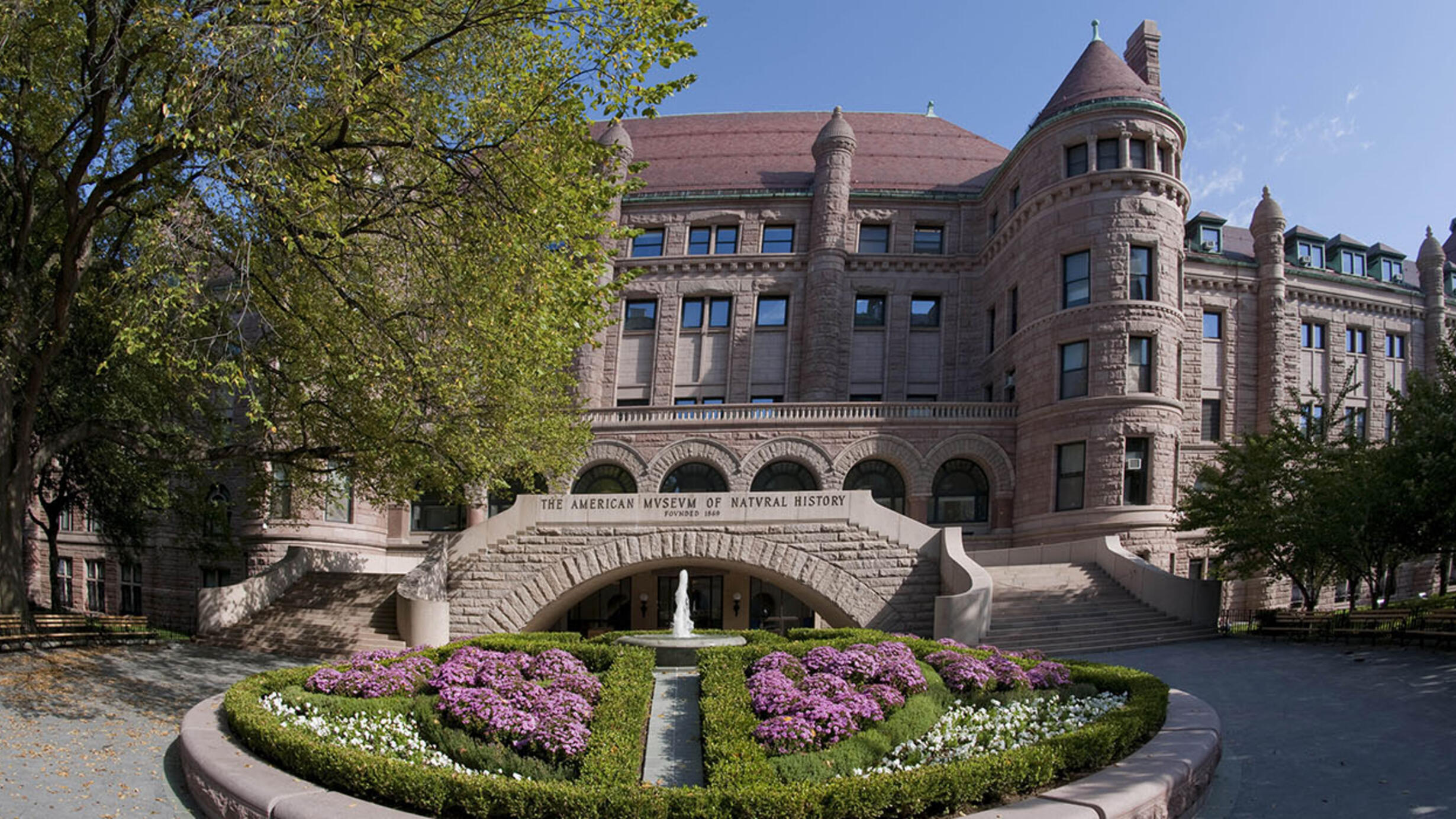 Circular fountain surrounded by flowers in front of the 77th street facade of the American Museum of Natural History.