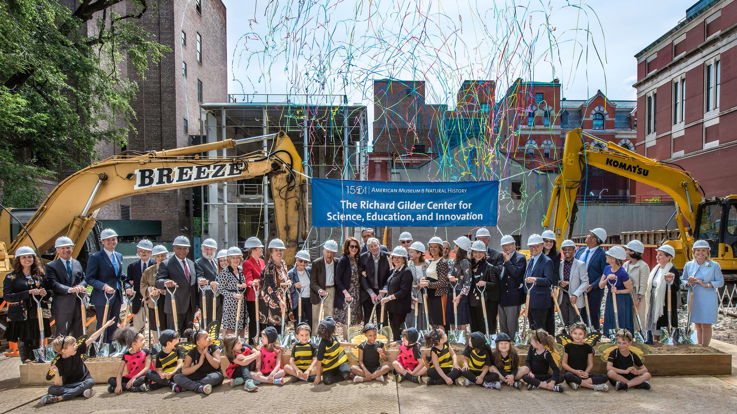 Group of people wearing hard hats and holding shovels, stand behind a group of seated children to commemorate the Gilder Center groundbreaking.