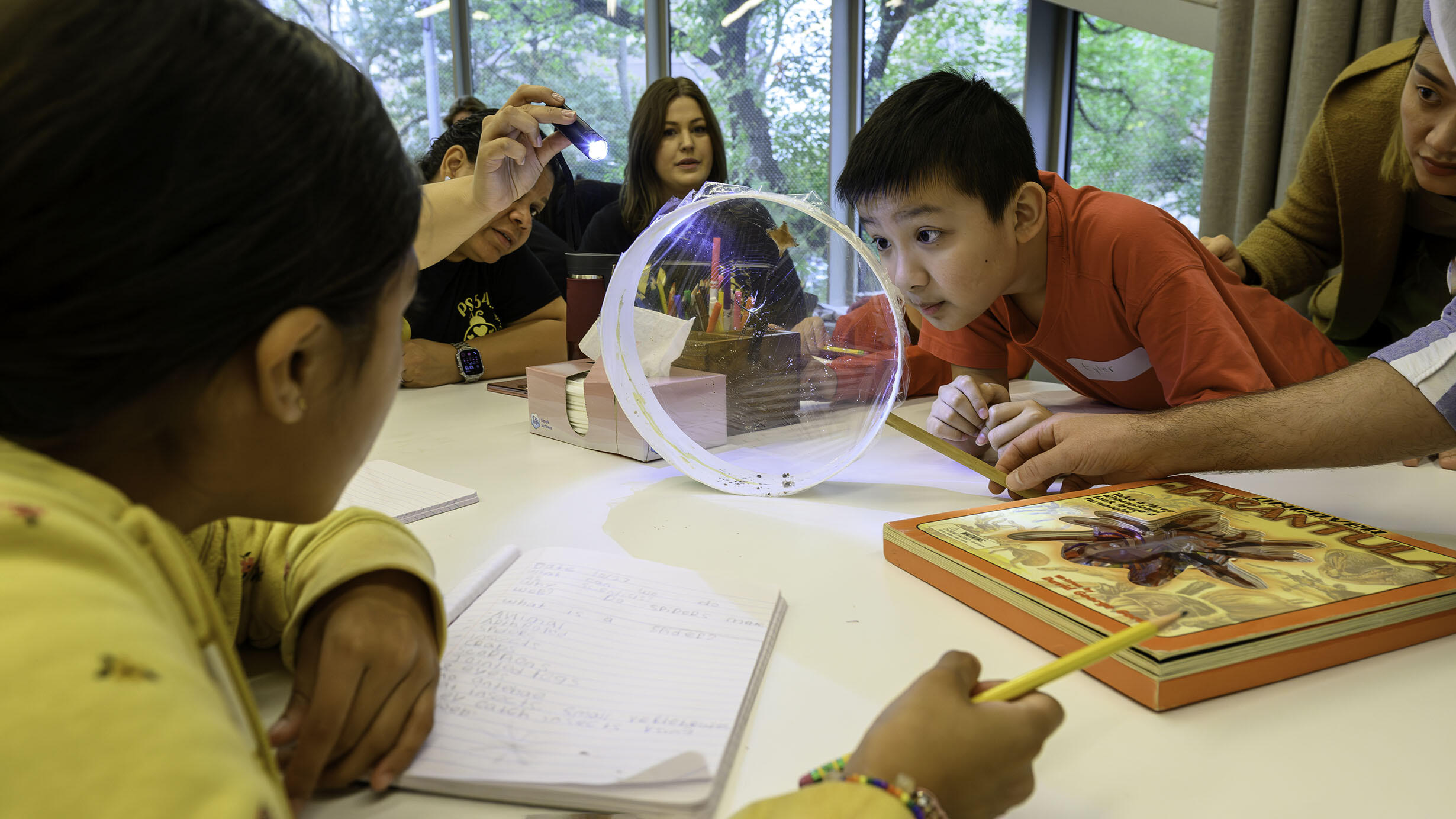 Two young students and three adults sit around a large table observe a spider in a round container at the center of the table.