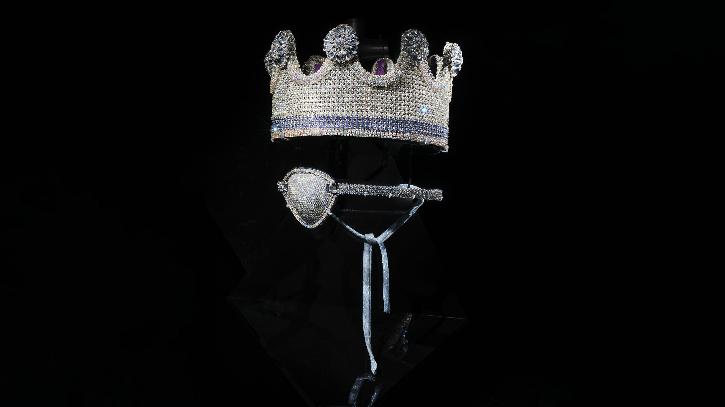 A jewel encrusted crown and eyepatch, featuring platinum and diamonds, against a dark background.