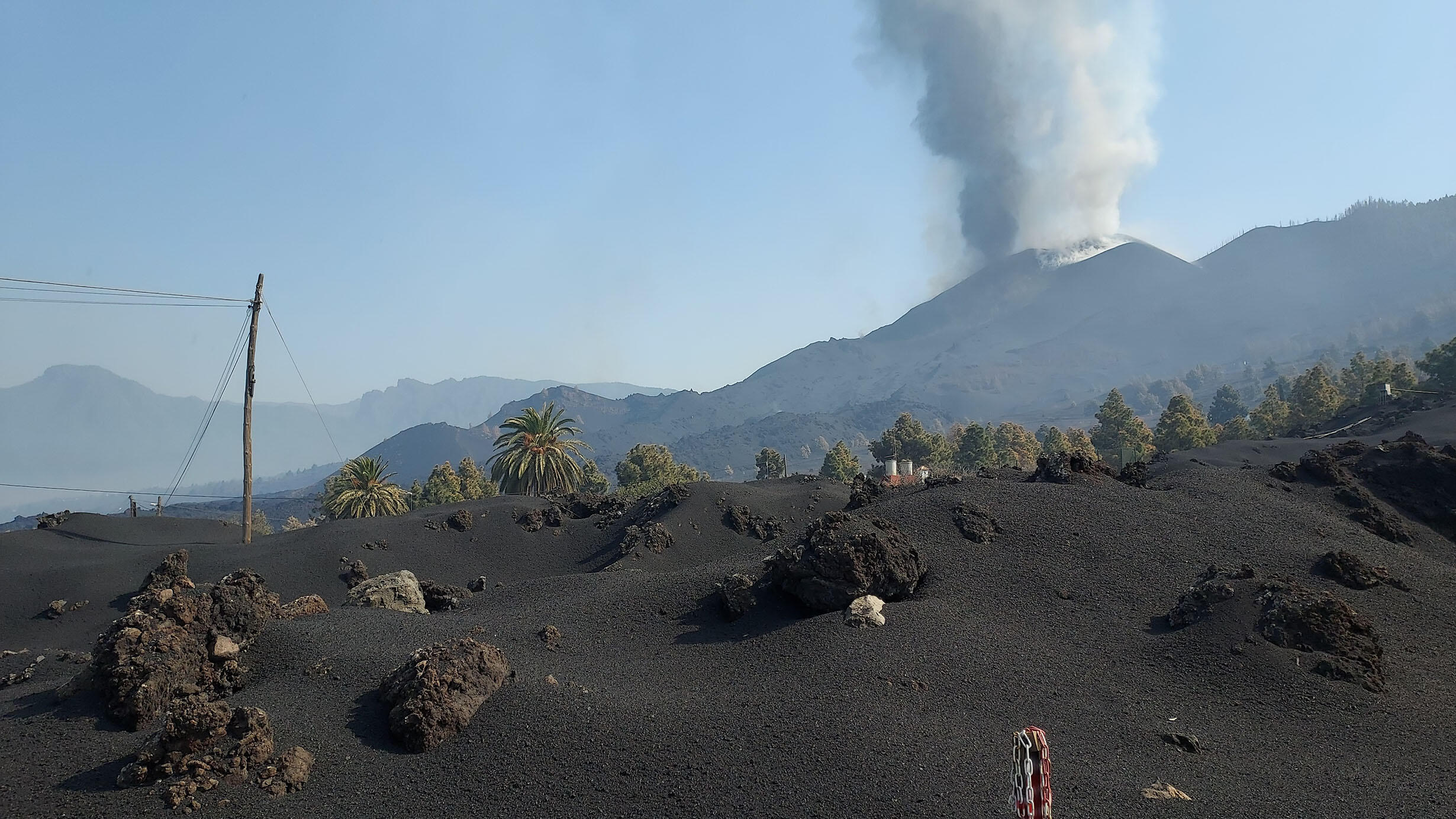 A plume of smoke emerges from a volcano in the distance past hills covered in volcanic ash and trees.