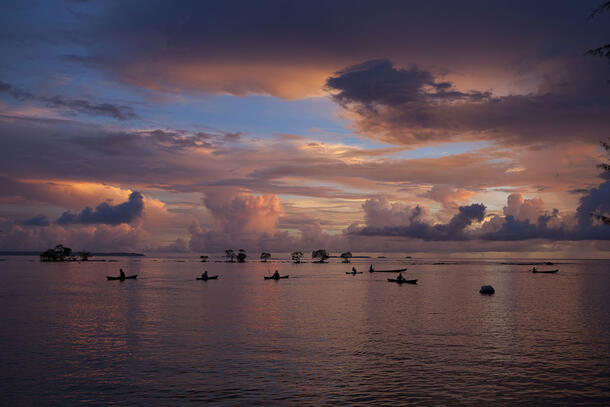 A wide swatch of water with the silhouettes of seven boats and fishers. The sky is filled with pink and purple clouds.