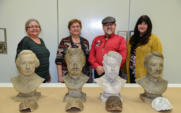 Four people pose behind a row of plaster cast busts of four people on a table.