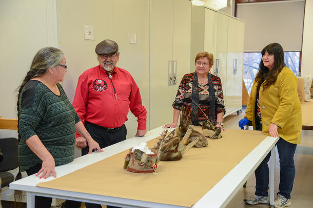 Four people stand around a long table. On the table are four headpieces made with fur, feathers, and animal skin.