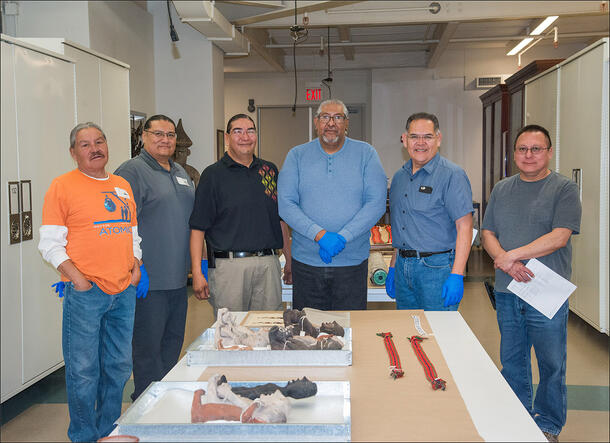 Six people pose in a row and stand in front of a table with six figurine artifacts and additional items laid out on trays.