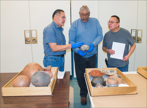 Mark Mitchell, Bernard Mora, and Larry Samuel examine a decorated Tesuque pottery bowl behind two tables holding wooden trays full of pottery pieces.