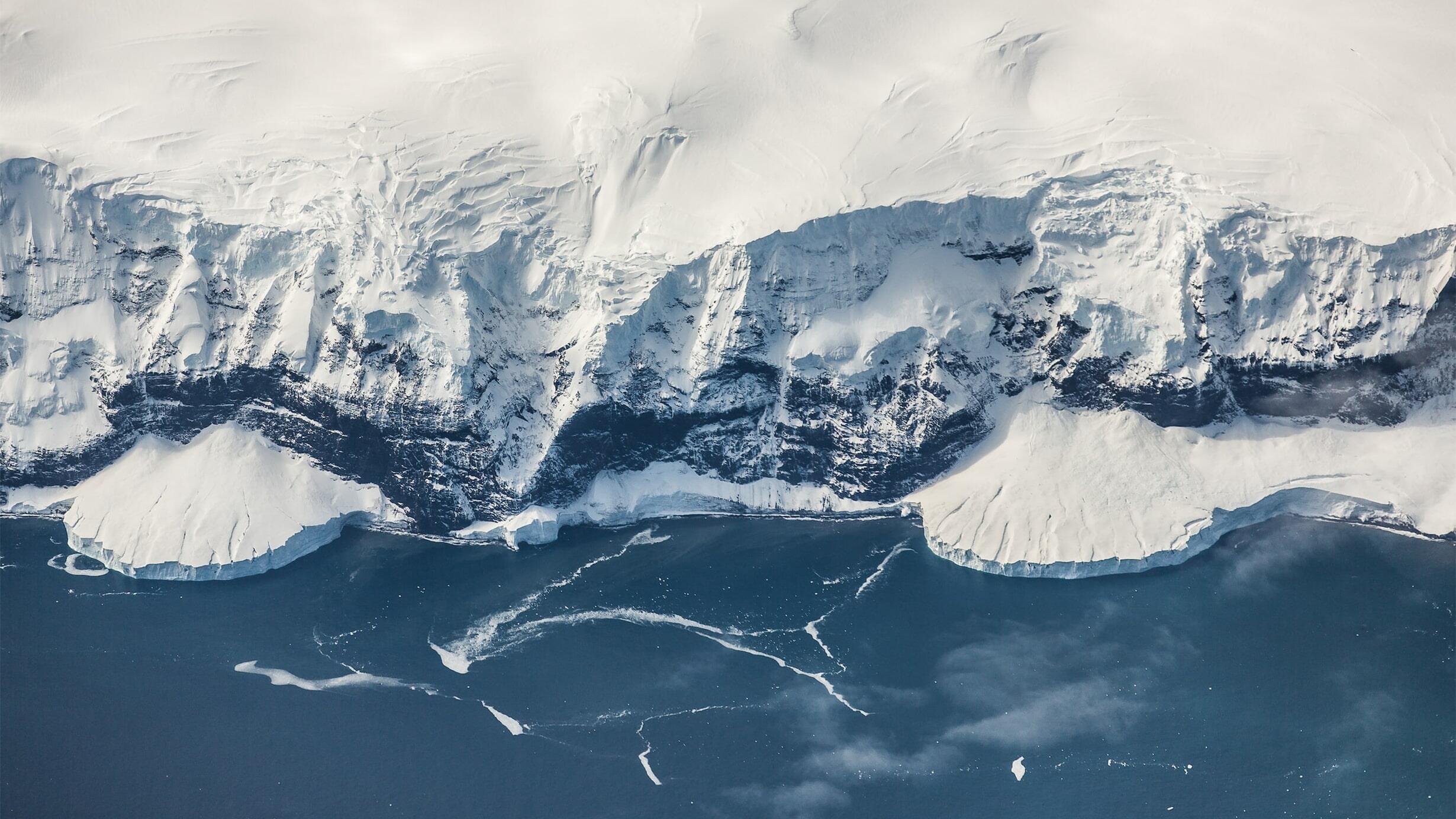 An aerial photograph of glacial ice at the edge of a small island, off the coast of Antarctica, meeting the Southern Ocean.