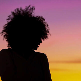 A woman's face, silhouetted against the sky, her curly hair outlined against purple and red hues of a sunset.