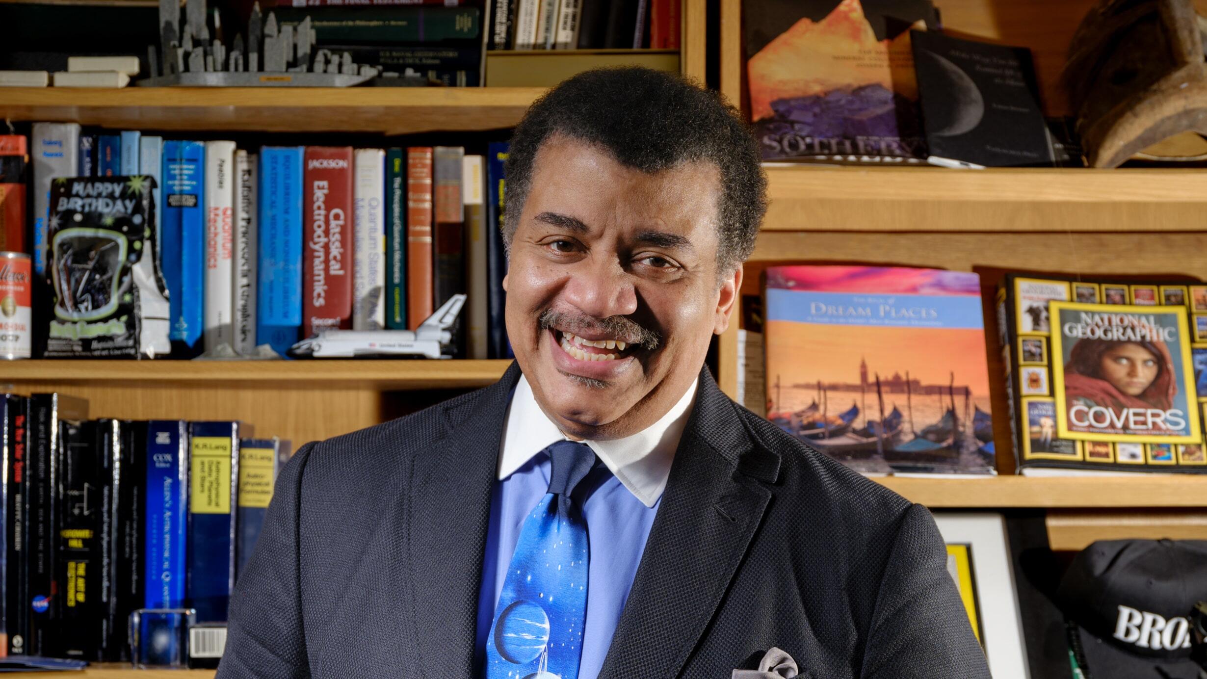 A portrait of astrophysicist Neil deGrasse Tyson, seen wearing a colorful tie depicting the solar system, shelves of books in the background.