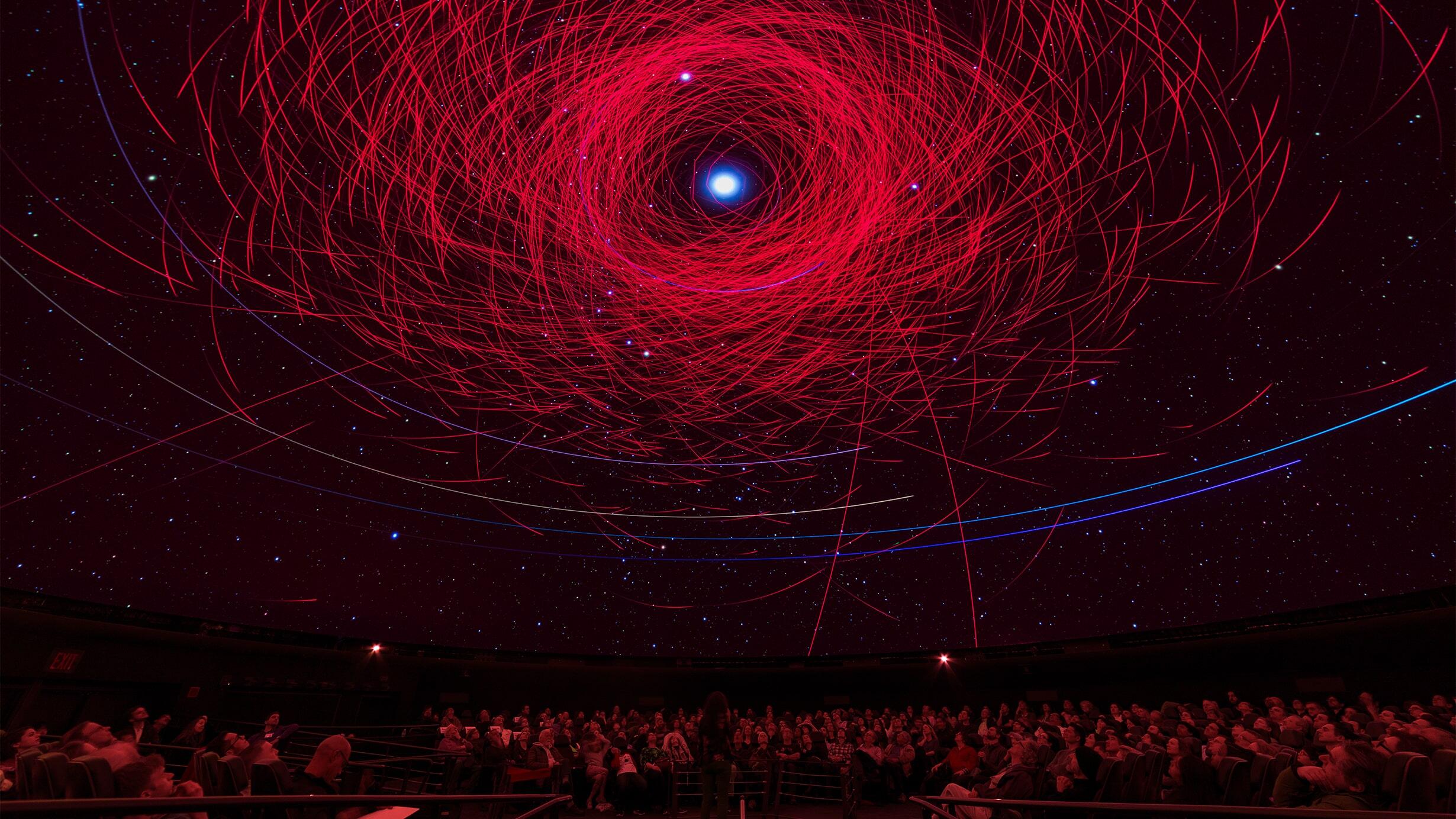 A crowd inside the Hayden Planetarium gazes up at bright, sharp lines depicting the paths of objects in our solar system.