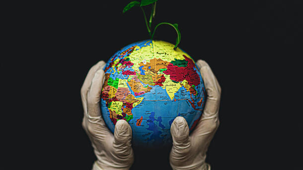 A pair of gloved hands hold up a small globe that represents earth and has a few leaves sprouting from the top.