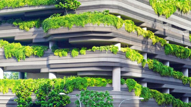 A multi-story, modern concrete building, with sleek horizontal lines, and generous amounts of shrubbery flowing from multiple balconies.