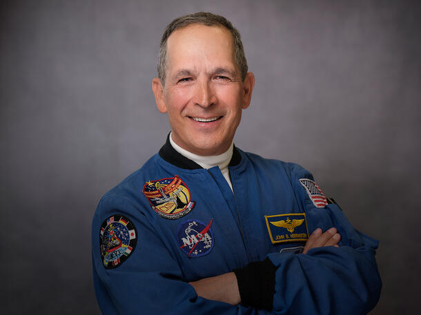 Smiling portrait of John Herrington, standing with arms crossed and wearing a jacket with multiple patches, with his name, NASA, and an american flag.