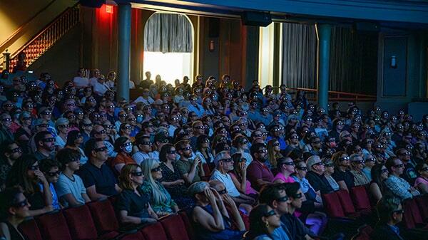An audience in a theater, its attendees wearing 3D glasses as they face the screen.