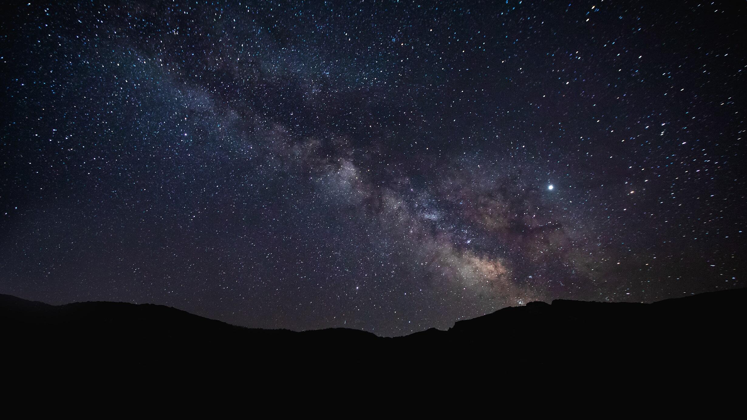 A panoramic view of a mountain range against a backdrop of the night sky, with bright stars and colored splotches of the Milky Way.