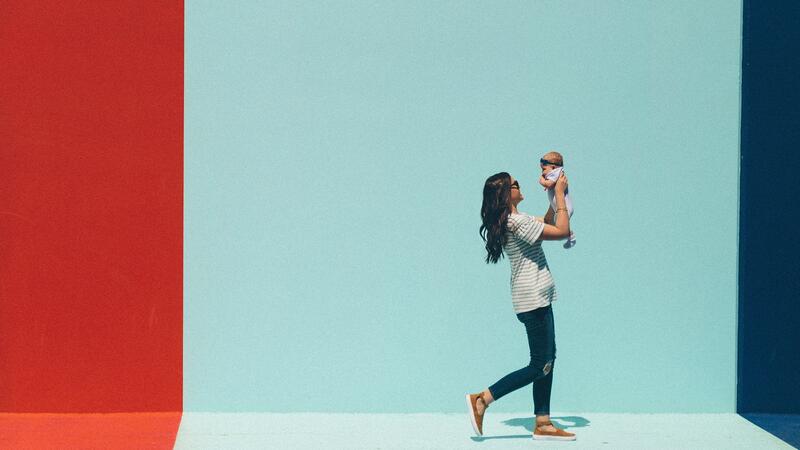 A woman walking, holding her infant in the air, on a bright day.