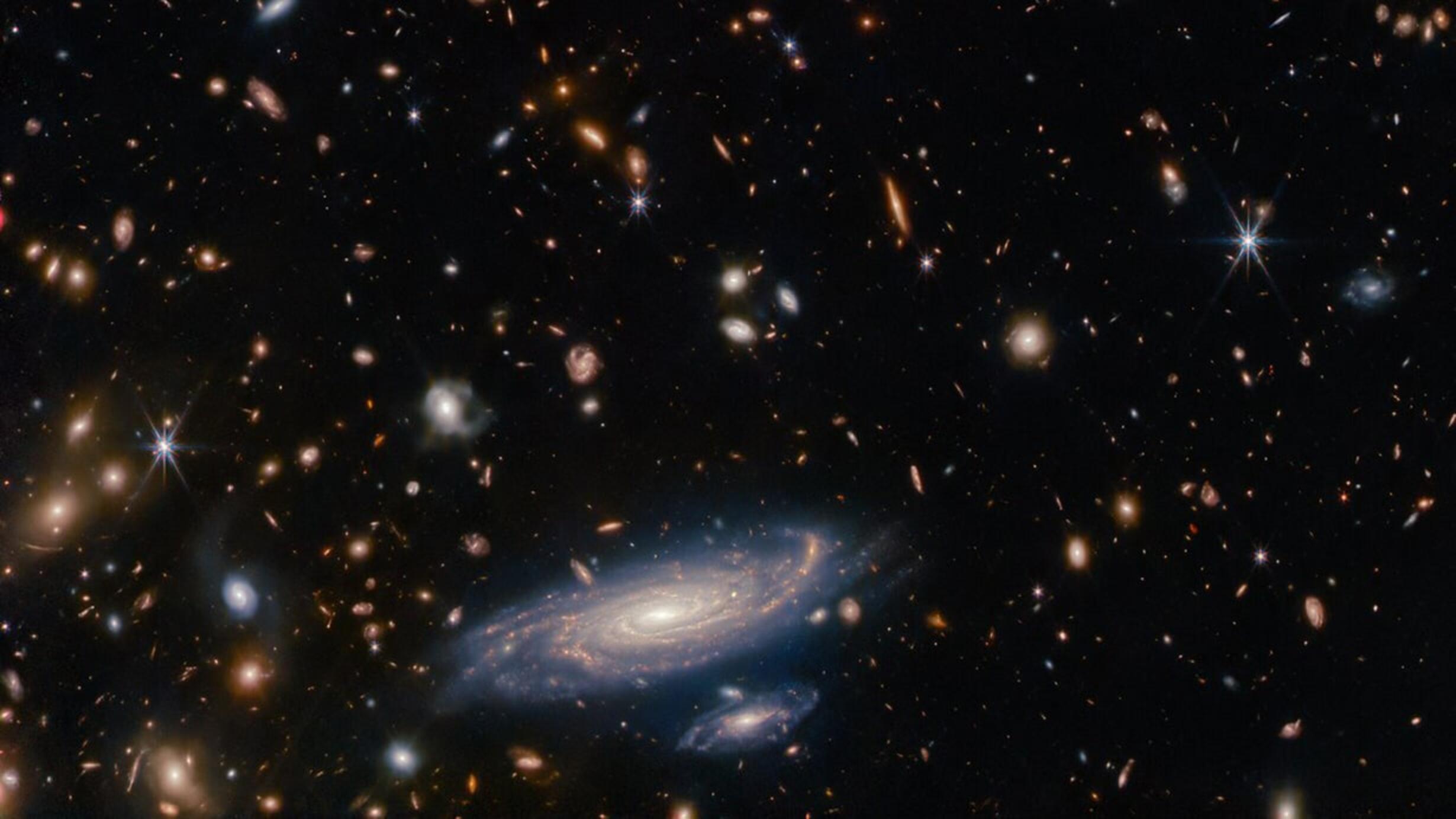 Many stars and galaxies lie on a dark background, in a variety of colours but mostly shades of orange. Some are large enough to make out spiral arms.