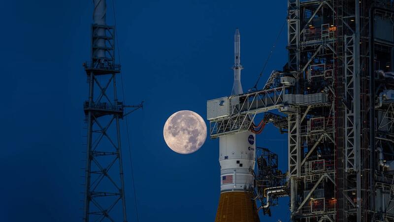 NASA's Orion spacecraft, its nosecone pointing skyward, seen against a deep blue night sky with a vivid full moon.