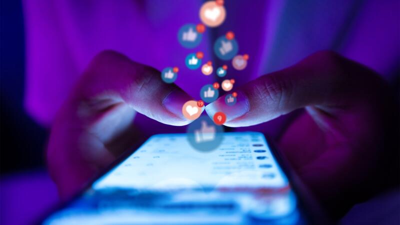 Two thumbs in close up, tapping on a bright phone screen. Thumbs up and heart emojis float above, depicting the likes and loves of social media.