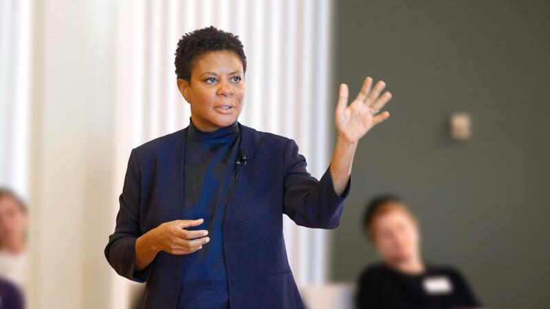 Alondra Nelson, in a dark blue suit against a white curtain, standing and addressing a seated audience.