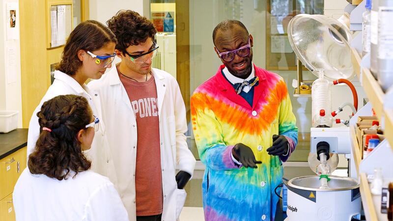 Dr. Andre Isaacs, wearing a bright rainbow tie-dyed lab coat, demonstrating the use of equipment to three young lab assistants.