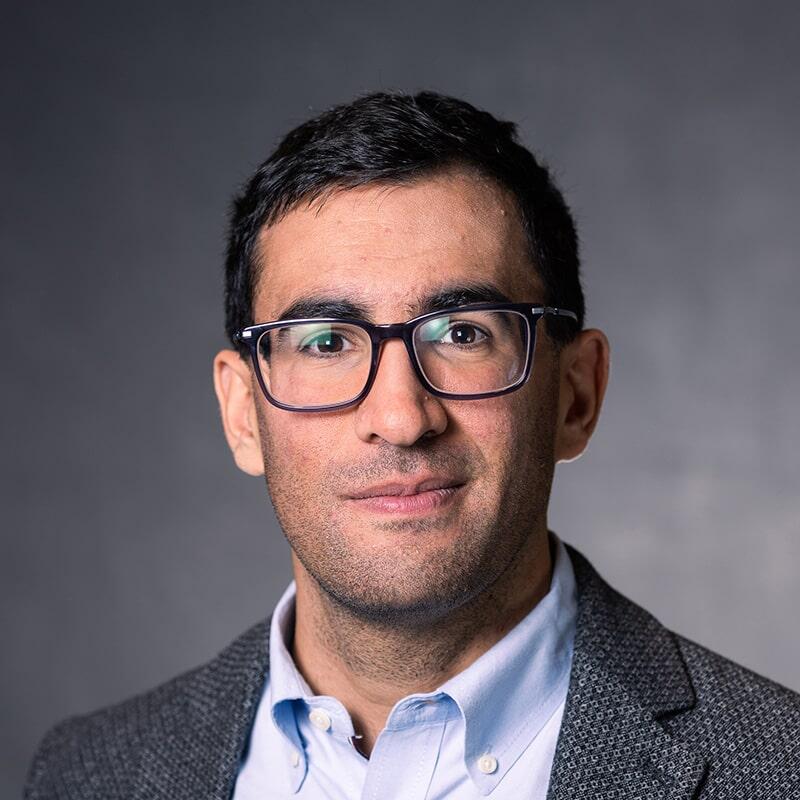 A close-up photograph of Daniele Visioni, wearing a light collared shirt and a gray sportcoat, against a gray background.