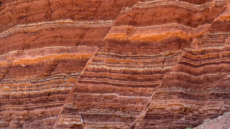 A sheer wall of reddish rock in the desert. Clearly delineated, horizontal layers with distinct coloration correlate with different geological eras.