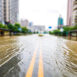 A city street in China is flooded - water cascades over the paved urban road, with tall buildings in the background.