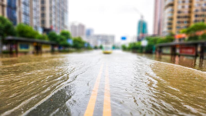 A city street in China is flooded - water cascades over the paved urban road, with tall buildings in the background.