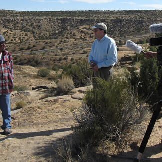 Harold Polingyumptewa and Peter Whiteley stand facing one another on top of rocky ridge, filmed by two camera men.