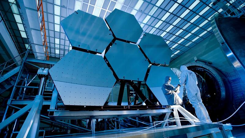 Six large hexagonal mirrors, part of the James Webb Space Telescope, being inspected by a NASA scientist in a large facility.