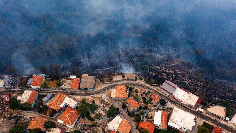An urban wildfire, seen from above. Red terra cotta rooftops, untouched by flame, sit across from the smoky ruins of burnt homes.