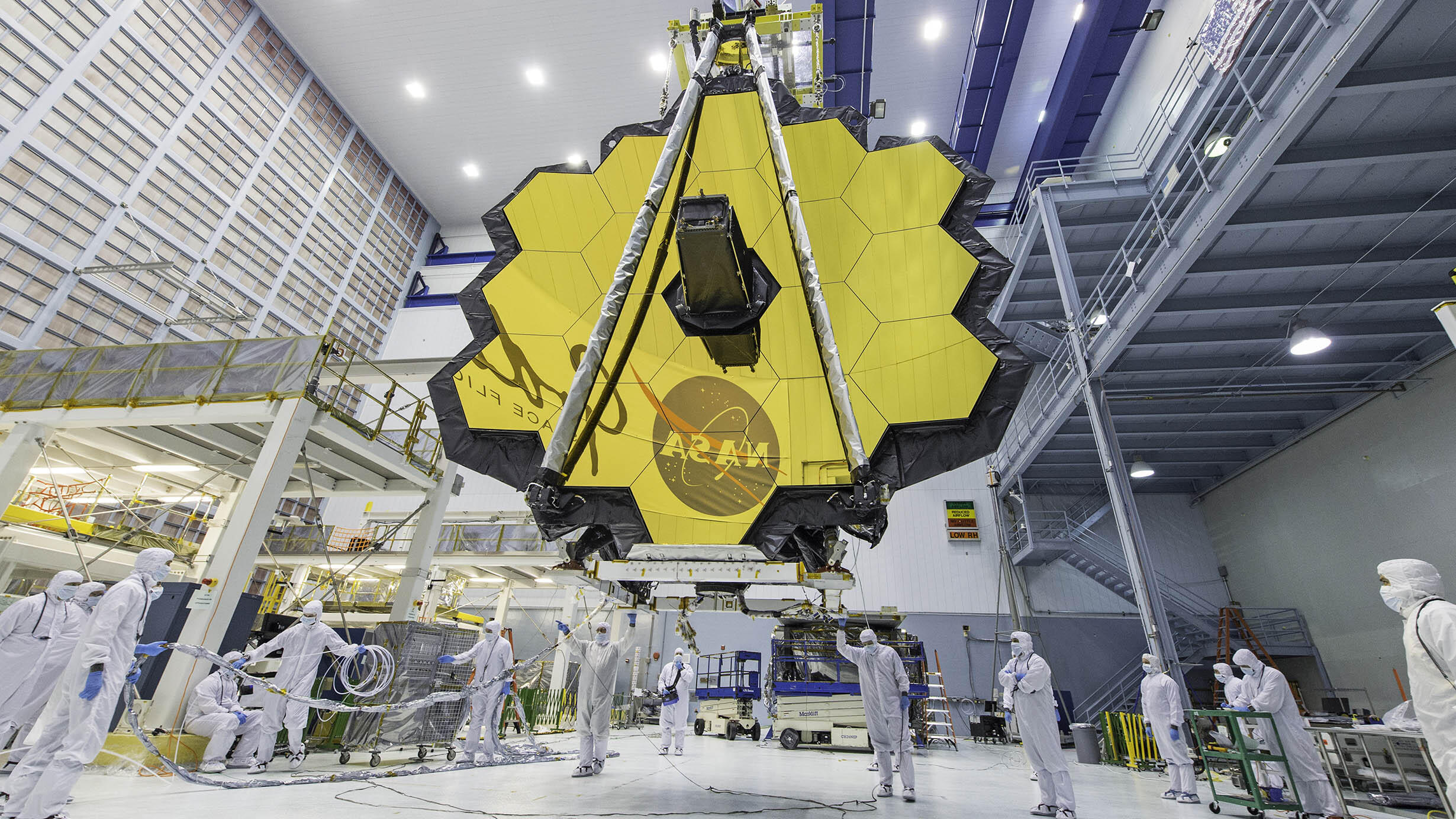 Fourteen technicians wearing cleanroom suits stand beneath the James Webb Space Telescope.
